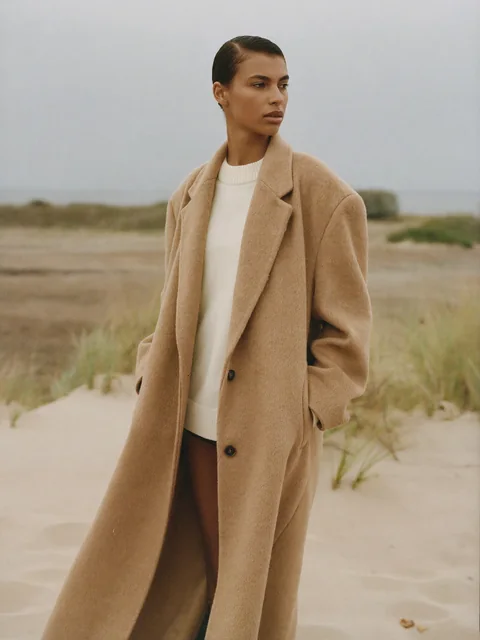 VEYRA Shoreline: the long camel coat over a cream knit, among the dune grass