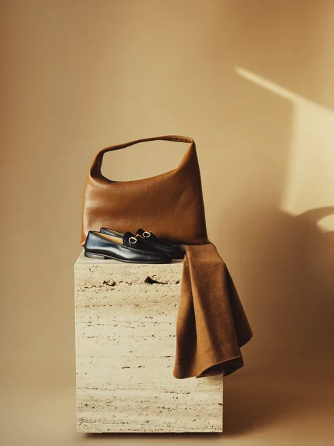 A cognac leather bag and black loafers on a travertine plinth